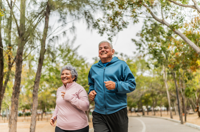 Older man and woman happily walk outside along a tree-lined path.