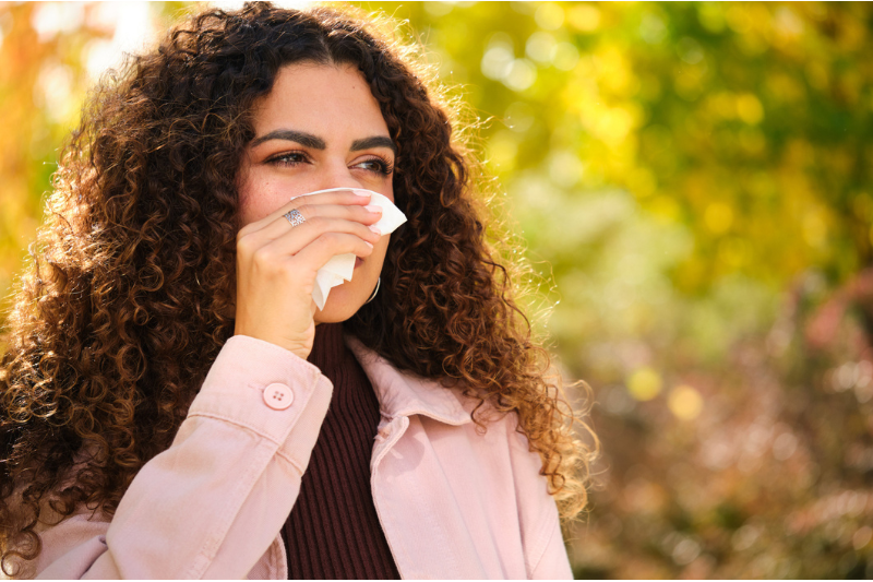 A young Caucasian woman with curly hair blows her nose into a tissue during autumn.