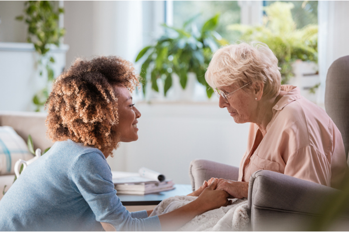 African American female home nurse talks with a senior woman, sitting in the living room and holding her hands.