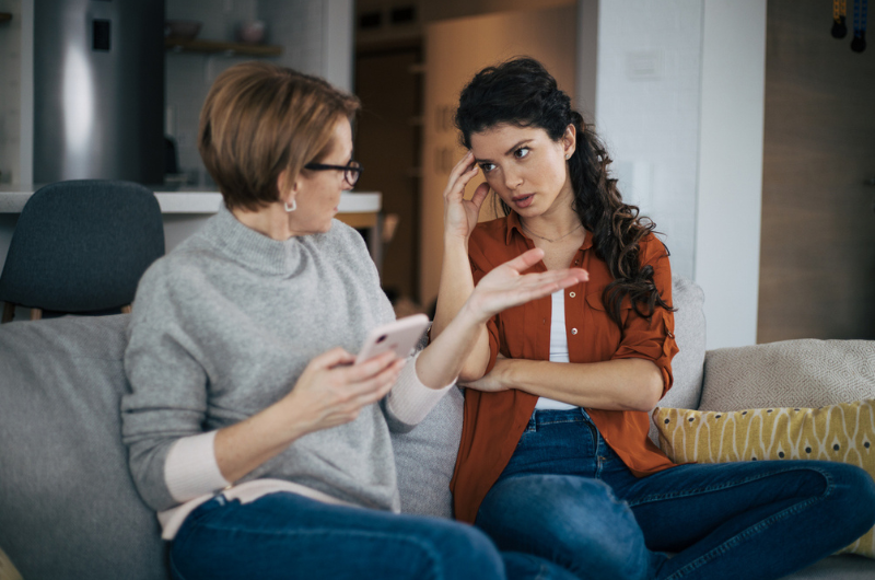 Brunette woman sits on a couch, arguing with her baby boomer mom.