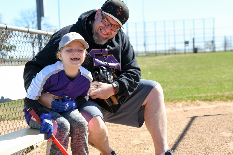 Russell and his daughter Devyn at the baseball field, sitting and smiling together.