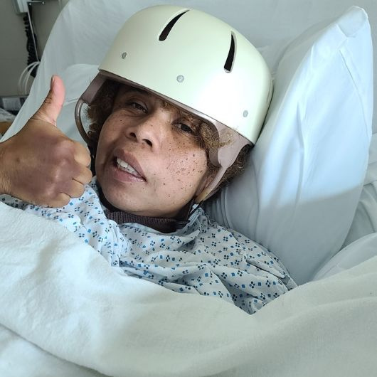 Aretha in the hospital, smiling in her bed while wearing a protective helmet after her brain surgery.