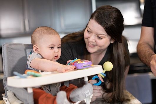 Baby Benny sitting in a high chair, playing with toys, with his mother smiling at him.
